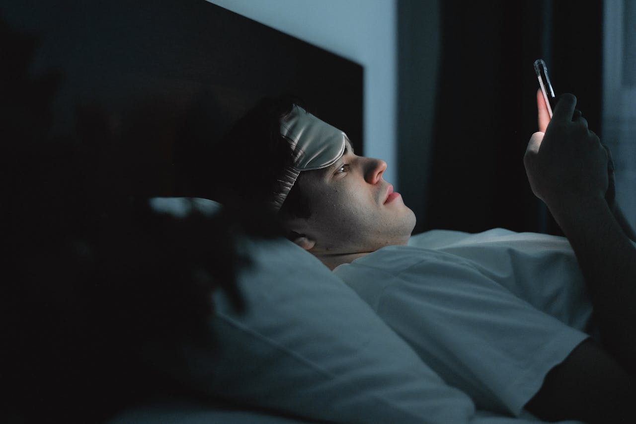 About A man wearing a sleep mask late at night using a smartphone on his bed.
